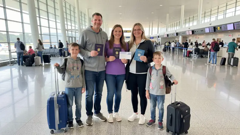 Family at airport holding passports and travel insurance card, parents with two children, rolling suitcases nearby